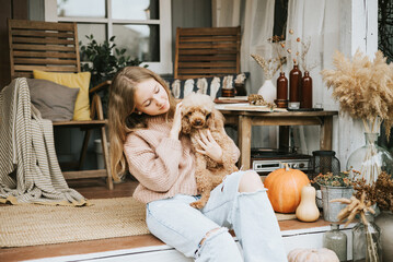 beautiful teenage girl in cozy warm knitted sweater sits and hugs with poodle dog on porch of...