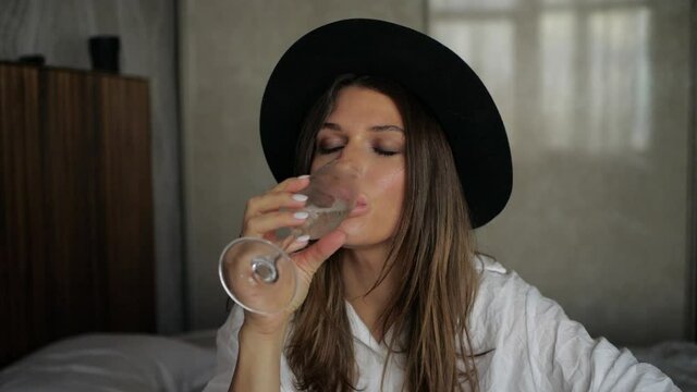 a beautiful girl in a hat and a white shirt seductively drinking champagne sitting on the bed in the hotel. High quality FullHD footage
