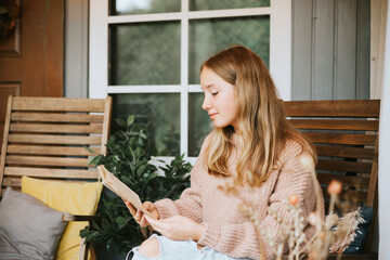 beautiful teenage girl in cozy warm knitted sweater and jeans is sitting on porch of house with cozy autumn decor and reading book, autumn season concept in pastel colors