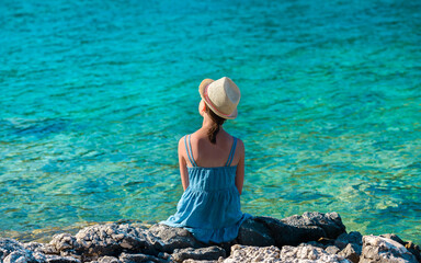 Little girl in a straw hat and a blue summer dress sitting on a rocky sea shore - a shot from behind her back