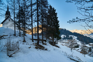 Alpejski kościółek wysoko w górach schowany za kępą drzew. Zimowy krajobraz górski.