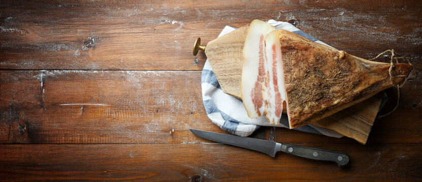 Bacon, guanciale, with cutting board and knife on wooden background, top view, space for text.