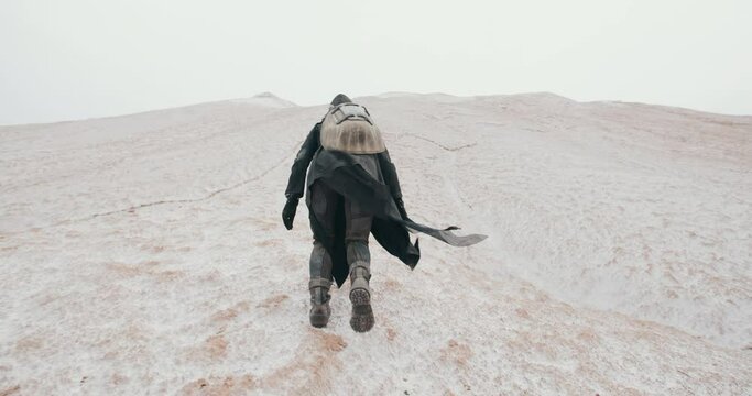 Backview wide shot post apocalyptic woman in grunge clothes hooded black mantle protective gas mask respiratory walking up hill in white desert. Cyberpunk soldier. Future female survivor with back
