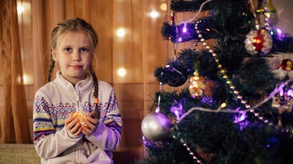 A girl with brown hair cleans a tangerine sitting near a decorated Christmas tree
