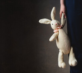 Cropped image of child's hand holding stuffed rabbit against dark wall - Powered by Adobe