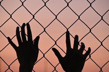 Front view silhouette of hands on a fence, sunset background.