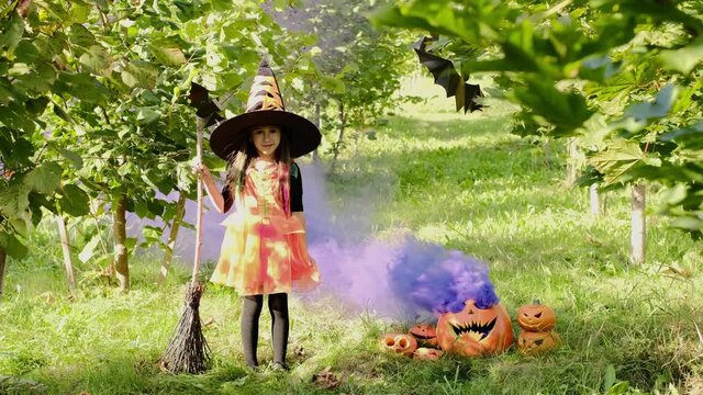 A girl in a witch costume with a broomstick stands among maple trees and colored purple smoke coming from a large pumpkin with a carved evil smile. halloween. 