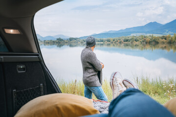 Car trunk view with woman legs in white sneakers on the man dressed warm knitted clothes and jeans enjoying the mountain lake view. Cozy early autumn couple auto traveling concept image.
