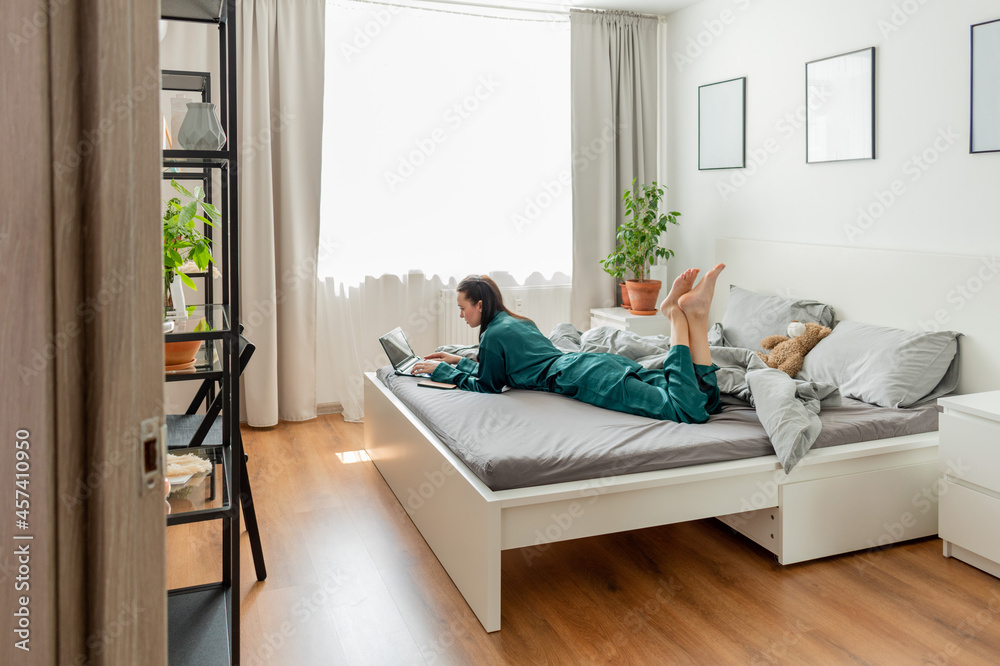 Wall mural young woman work at laptop while sitting on bed in bedroom. concept of remote work. wellness and men