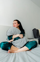 Obraz premium Young woman in silk pajamas sits on the bed and hugs a pillow. Loneliness inside the house. Wellness concept.