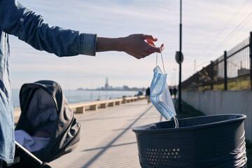 woman with baby carriage throwing her mask in the trash on a promenade, end of pandemic thanks to covid-19 vaccine, post-pandemic, freedom, virus-free holiday, virus-free summer