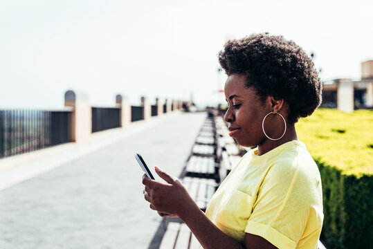 Portrait of black girl with afro hair and hoop earrings using her mobile phone in an urban space in the city.