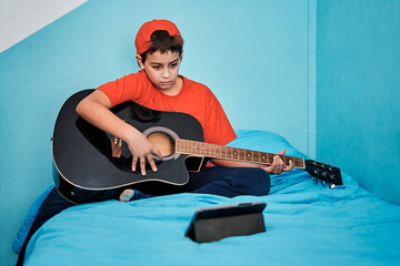 Boy learning to play guitar looking at tablet on top of bed in room