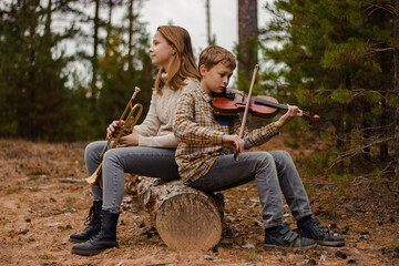 Boy and girl, brother and sister, teenagers play the trumpet and violin sitting in the woods on a log.