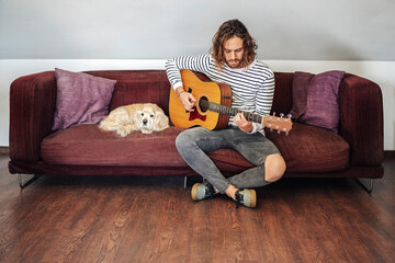 Attractive man with long hair playing acoustic guitar indoor with dog