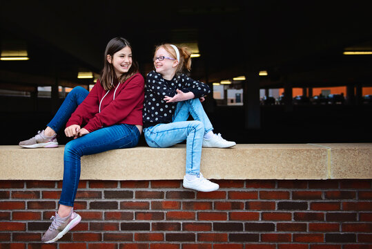 Two happy tween girls sitting on a brick wall together talking.