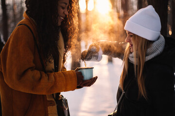 Happy woman pouring tea for girlfriend standing at park during winter