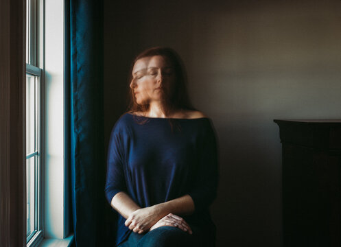 Woman with blurry face sitting alone in a dark room beside a window.