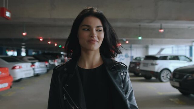 Following beautiful happy brunette woman wearing black leather jacket and sunglasses walking along underground parking with parked looking to camera flirts and smiling. Urban style fashion