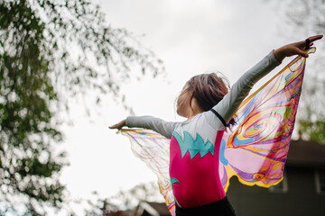 A little girl in transparent butterfly wings lifts arms towards sky