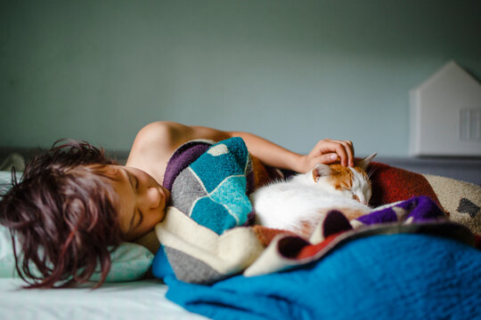 A child lays in bed under a pile of cozy blankets snuggling with a cat