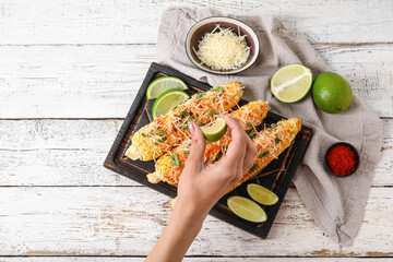 Woman squeezing lime onto tasty Elote Mexican Street Corn on white wooden background