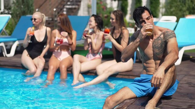 Confident Middle Eastern tattooed man drinking whiskey in slow motion resting at poolside in sunshine. Portrait of masculine handsome tourist satisfied with leisure at resort outdoors. Tourism and joy