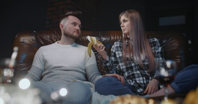 Millennial couple having fun watching TV. Young female eating banana, her boyfriend looks at her, flirts, takes condom and show it playfully offers sex to girlfriend. Concept of sexual relationships