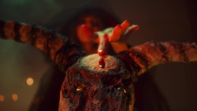 Crazy witch woman making fingerprint with blood on cow skull, sitting in pentagram circle. Evil sorceress doing rite and sacrifices at night, using black witchcraft, candles on floor. Halloween time.