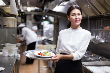 Successful waitress standing in restaurant kitchen with ordered meals, ready to serving guests