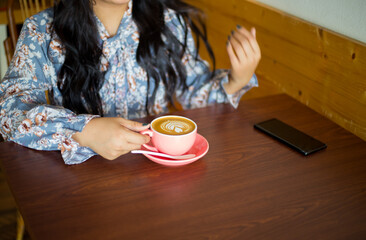 A young girl in a cafe sitting and holding a cup of coffee to drink. Unrecognizable young woman holding tasty coffee latte in the morning. 