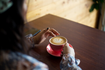 Horizontal closeup shot of a young woman sitting in a café holding a cup of latte. Indoor lifestyle portrait of unrecognizable girl holding beautifully decorated latte art coffee. 