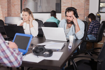 Portrait of bearded Spanish man in headphones working on laptop in busy modern coworking office