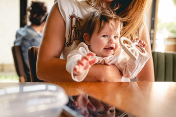 Close up on small happy caucasian baby girl or boy infant smiling in hands of her mother sitting by the table at restaurant or cafe in bright day childhood and growing up concept real people