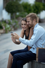 Smiling young couple resting on bench in park and watching funny videos on smartphone