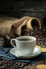 Hot coffee in a white coffee cup and many coffee beans placed around and sugar on a wooden table in a warm, light atmosphere, on dark background.