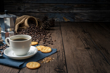 Hot coffee in a white coffee cup and many coffee beans placed around and sugar on a wooden table in a warm, light atmosphere, on dark background.