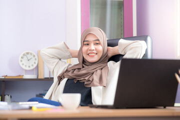 Asian Muslim woman office worker sitting in front of a laptop computer at a desk and smiling...