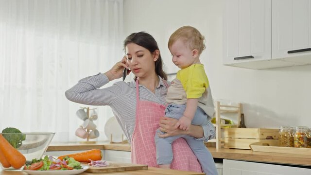 Caucasian busy mother doing housework with baby boy toddler in kitchen. Beautiful single mom use phone call for work and cook foods prepare breakfast for little kid son in house. Family-housekeeping.