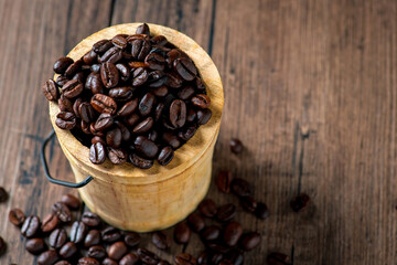 A small wooden bucket of coffee and many coffee beans were placed around on a wooden table in a warm, light atmosphere, on a dark background, with copy space.