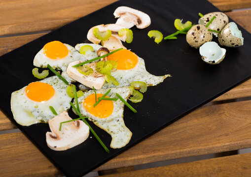 Delicious broken quail eggs with green onion and sliced champignons on black serving board
