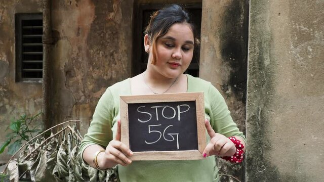 A young girl holding a slate with the Stop 5G  written on it for people. Medium shot of an Indian female showing a blackboard in casual dress.Concept of Outdoor portrait of Caucasian young woman.