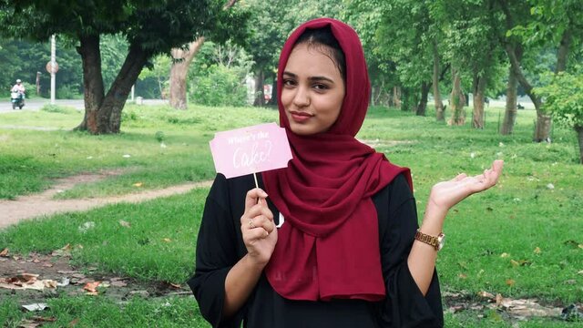 Young Afghan women posing in hijab using pink photo booth props written where is the cake.Young happy woman holds a paper on a stick and shows finger towards stick, standing next to a lake.