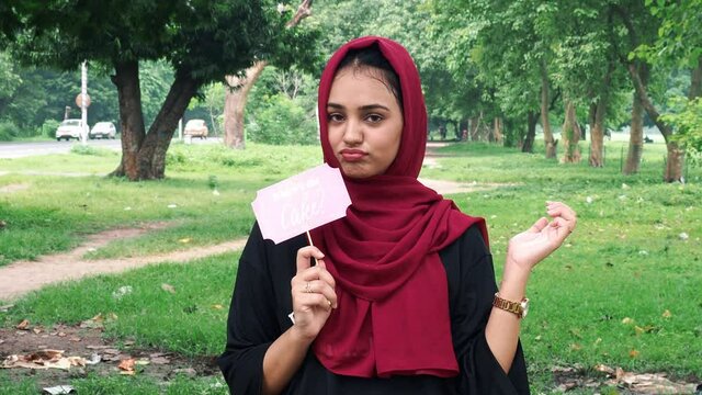 Young Afghan women posing in hijab using pink photo booth props written where is the cake.Young happy woman holds a paper on a stick and shows finger towards stick, standing next to a lake.outdoor.