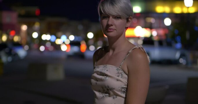 Cute and confused girl women wearing summer dress standing in front of bokeh bubble lights and passing cars at night, in dark dramatic lighting. In Cinema 4K (4096x2160) 30fps slowed from 60fps.