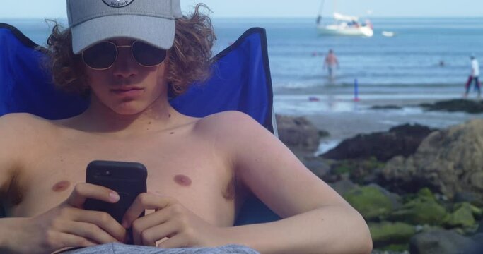 Curly-haired Man Sitting And Using Smartphone At Coverack Beach In Cornwall, UK. static