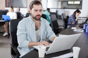 Portrait of bearded young man working on laptop in busy modern coworking office