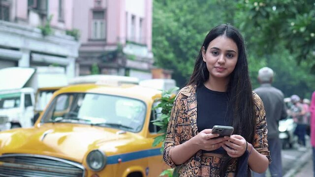 Kolkata, India : A young woman standing in front of yellow taxi in Kolkata talks on the phone and takes a photo with a smartphone in her hands. Summer sunny day in the city