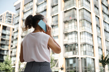 View from the back of a young African American mixed race woman talking on mobile phone on the background of urban high buildings. Business and communication concepts