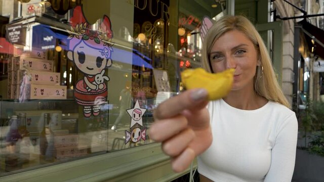smiling blond woman eats sweet french macaron, offers bite to camera